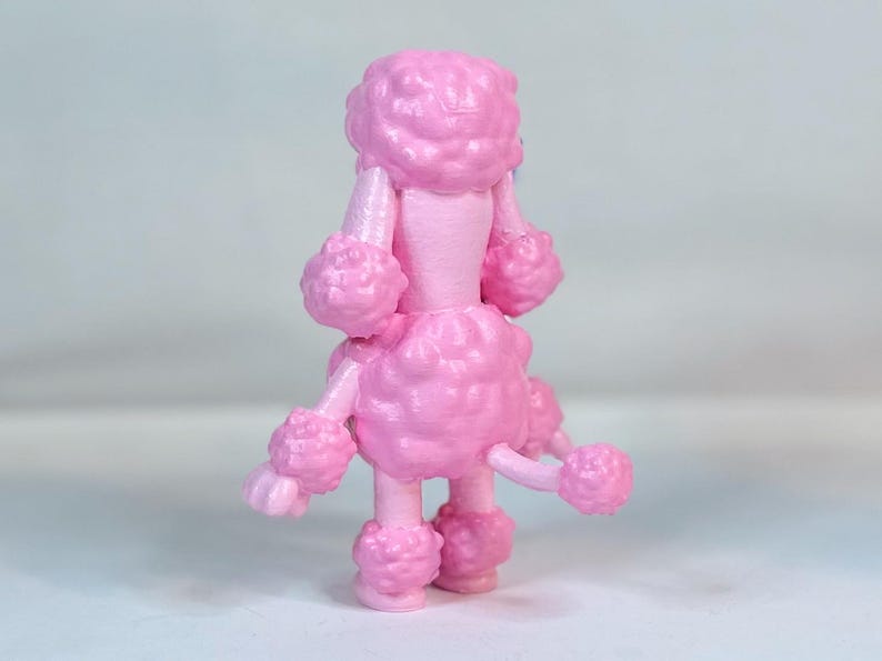 K&ouml;nnte beinhalten: Eine rosafarbene Pudelfigur mit einem flauschigen, lockigen Fell. Der Pudel steht auf einer wei&szlig;en Oberfl&auml;che.
