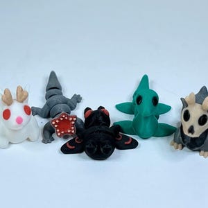 May include: Five small, 3D printed figurines of different creatures. A white creature with red eyes and antlers, a gray lizard-like creature, a black creature with red eyes and wings, a green creature with green eyes, and a gray creature with a skull-like face and antlers.