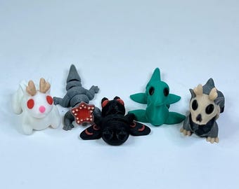 May include: Five small, 3D printed figurines of different creatures. A white creature with red eyes and antlers, a gray lizard-like creature, a black creature with red eyes and wings, a green creature with green eyes, and a gray creature with a skull-like face and antlers.