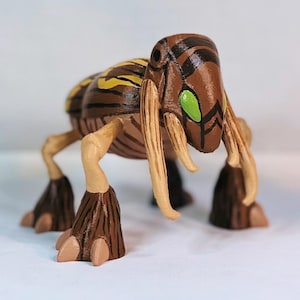 Peut inclure: Une figurine imprimée en 3D peinte en brun et jaune représentant une créature avec de longues pattes fines, un corps segmenté et deux grands yeux verts. La créature a deux longues cornes fines de couleur ivoire.