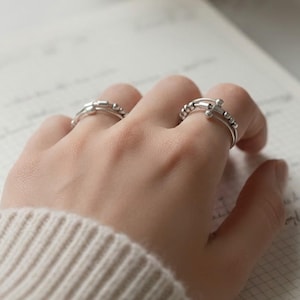 Puede incluir: Primer plano de dos anillos de plata en una mano, con un tercer anillo visible en el fondo. Los anillos tienen un diseño minimalista, con pequeñas cuentas y una banda sencilla. La mano descansa sobre un cuaderno abierto con notas manuscritas.