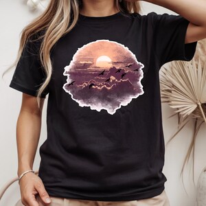 以下が含まれることがあります： 夕焼けと飛んでいる鳥の水彩デザインが施された黒いTシャツ。アートワークには、オレンジ、紫、黒の色合いの太陽、雲、鳥が含まれています。このシャツは、クラシックなクルーネックです。