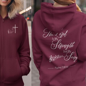 Könnte beinhalten: Burgunderfarbener Kapuzenpullover mit Reißverschluss. Auf der Vorderseite befindet sich ein weißes Kreuz und ein Blumendesign. Auf der Rückseite steht der Text "The Lord Is My Strength And My Song", Noten und die Bibelstelle Psalm 118:14.