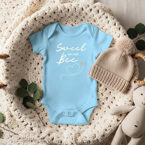 Puede incluir: Un body de beb&eacute; azul claro con el texto "Sweet as can Bee" en escritura blanca. Se incluye un peque&ntilde;o gr&aacute;fico de abeja amarilla. Un gorro de punto beige con un pomp&oacute;n y un oso de peluche tambi&eacute;n est&aacute;n en la imagen. Los art&iacute;culos est&aacute;n dispuestos en una cesta tejida.