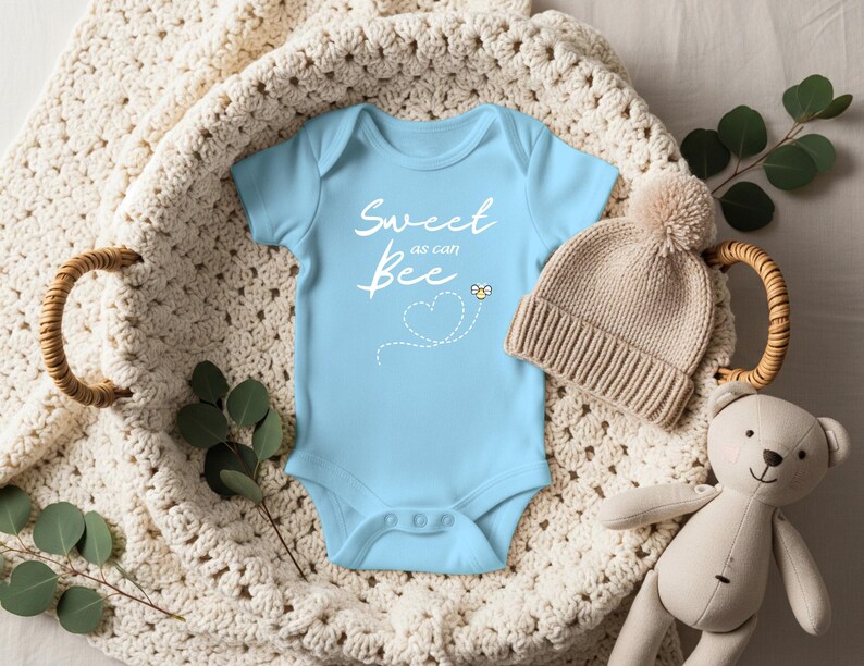 Puede incluir: Un body de beb&eacute; azul claro con el texto "Sweet as can Bee" en escritura blanca. Se incluye un peque&ntilde;o gr&aacute;fico de abeja amarilla. Un gorro de punto beige con un pomp&oacute;n y un oso de peluche tambi&eacute;n est&aacute;n en la imagen. Los art&iacute;culos est&aacute;n dispuestos en una cesta tejida.