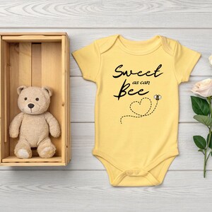 Puede incluir: Un body de beb&eacute; amarillo con el texto "Sweet as can Bee" y un gr&aacute;fico de abeja. Un oso de peluche est&aacute; en una caja de madera. Una rosa con dos flores est&aacute; a la derecha.