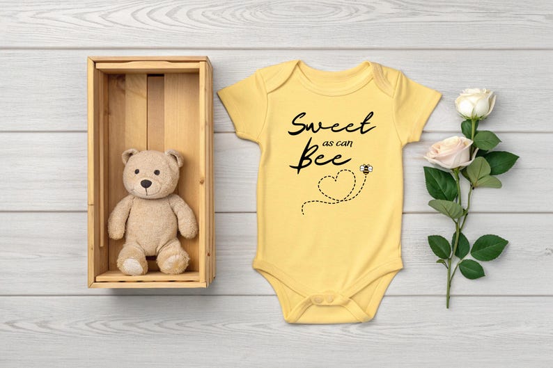 Puede incluir: Un body de beb&eacute; amarillo con el texto "Sweet as can Bee" y un gr&aacute;fico de abeja. Un oso de peluche est&aacute; en una caja de madera. Una rosa con dos flores est&aacute; a la derecha.