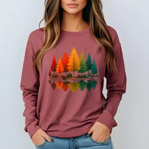 May include: A long-sleeved maroon shirt featuring a colorful graphic of stylized trees in shades of red, orange, yellow, and green. The trees are reflected in a body of water, creating a symmetrical design. The shirt is made of a soft material.