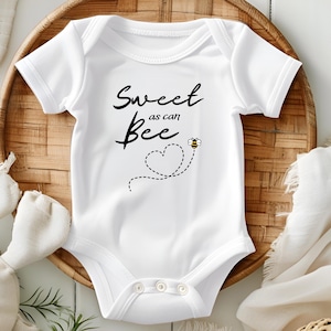 Puede incluir: Body de beb&eacute; blanco con el texto "Sweet as can Bee" en escritura negra. Un coraz&oacute;n punteado y un peque&ntilde;o gr&aacute;fico de abeja est&aacute;n debajo del texto. El body est&aacute; sobre una bandeja tejida con un peluche y un pa&ntilde;o blanco.