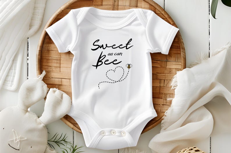 Puede incluir: Body de beb&eacute; blanco con el texto "Sweet as can Bee" en escritura negra. Un coraz&oacute;n punteado y un peque&ntilde;o gr&aacute;fico de abeja est&aacute;n debajo del texto. El body est&aacute; sobre una bandeja tejida con un peluche y un pa&ntilde;o blanco.