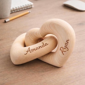 May include: Two interlocking wooden hearts, engraved with the names "Amanda" and "Ryan". The light-coloured wood has a smooth finish. The hearts are intertwined, symbolising connection and love. A thoughtful gift for couples.
