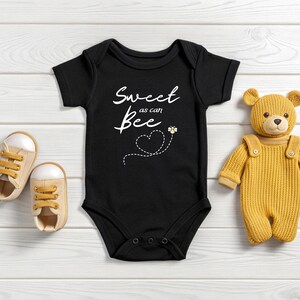 Puede incluir: Body de beb&eacute; negro con el texto "Sweet as can Bee" y un gr&aacute;fico de abeja. El body est&aacute; junto a un par de zapatos de beb&eacute; beige y un oso de peluche amarillo con peto.