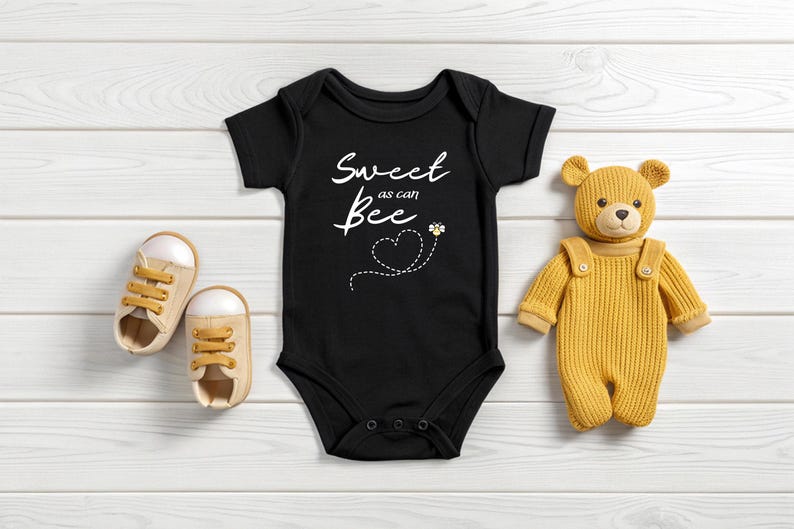 Puede incluir: Body de beb&eacute; negro con el texto "Sweet as can Bee" y un gr&aacute;fico de abeja. El body est&aacute; junto a un par de zapatos de beb&eacute; beige y un oso de peluche amarillo con peto.