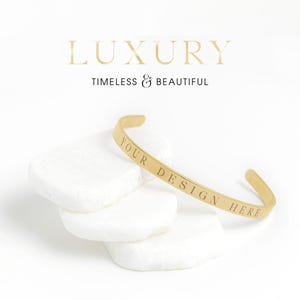 May include: A gold-coloured cuff bracelet with the engraved text "YOUR DESIGN HERE" is displayed on white, textured stones. The word "LUXURY" and the phrase "TIMELESS & BEAUTIFUL" are in the background.