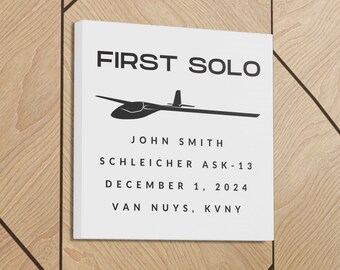 May include: A white canvas print with black text that reads "FIRST SOLO" and a black silhouette of a glider. The text below the glider reads "JOHN SMITH", "SCHLEICHER ASK-13", "DECEMBER 1, 2024", and "VAN NUYS, KVNY".
