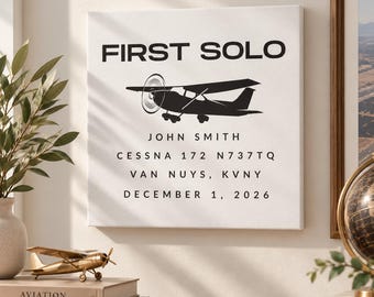 May include: A square white canvas wall art piece featuring black text and a black silhouette of a small airplane. The text reads FIRST SOLO, JOHN SMITH, CESSNA 172 N737TQ, VAN NUYS, KVNY, and DECEMBER 1, 2026, centered in a clean, sans-serif font.