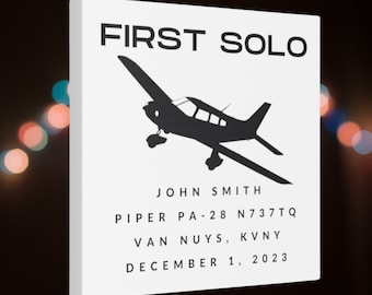 May include: White canvas art print with the text "FIRST SOLO" and a black silhouette of a Piper PA-28 airplane. Includes the name "JOHN SMITH", aircraft details, and the date December 1, 2023.