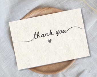 Puede incluir: Una tarjeta color crema con las palabras "thank you" en escritura elegante, acompañada de un pequeño corazón. La tarjeta descansa sobre un plato de madera, sobre un fondo de tela blanca y suave. El diseño es simple y elegante.
