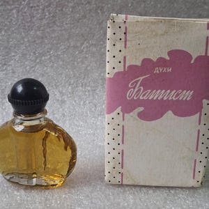May include: A vintage perfume bottle with a black cap and a clear, rounded glass body filled with amber liquid. The bottle is next to a rectangular box with the word "Baptist" in cursive on a pink cloud.