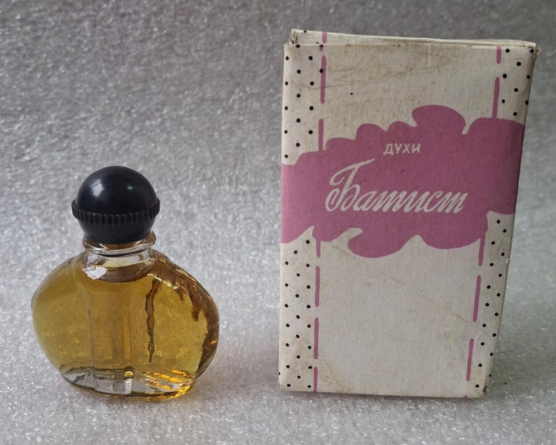 May include: A vintage perfume bottle with a black cap and a clear, rounded glass body filled with amber liquid. The bottle is next to a rectangular box with the word "Baptist" in cursive on a pink cloud.