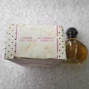 May include: A vintage perfume bottle with a black cap and a light brown liquid, next to a white box with black polka dots and text in Russian. The box has pink stripes and appears aged.