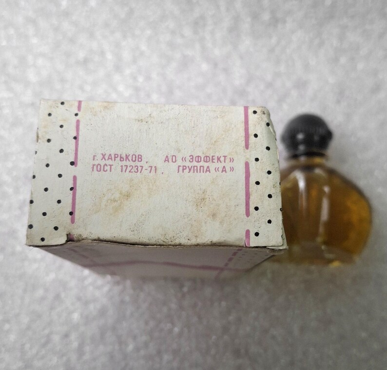 May include: A vintage perfume bottle with a black cap and a light brown liquid, next to a white box with black polka dots and text in Russian. The box has pink stripes and appears aged.