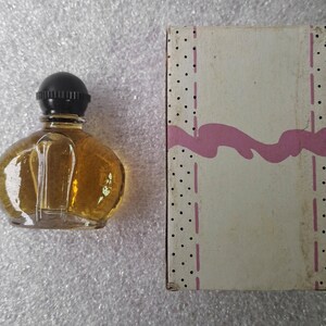 May include: A small, clear glass perfume bottle with a black cap, filled with amber liquid, sits next to a rectangular box. The box is off-white with a pink wavy design and black polka dots.