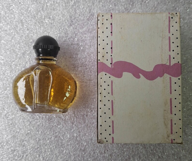 May include: A small, clear glass perfume bottle with a black cap, filled with amber liquid, sits next to a rectangular box. The box is off-white with a pink wavy design and black polka dots.