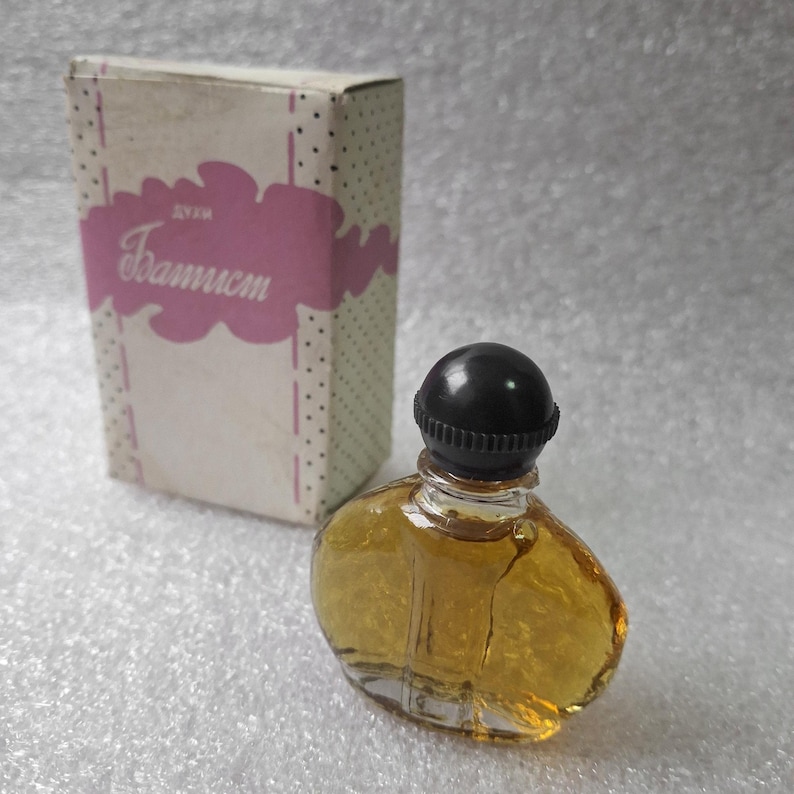 May include: A vintage perfume bottle with a black cap and a small, rounded glass body filled with amber liquid. The bottle sits next to a rectangular box with a pink label and the word "Baptist" in cursive.