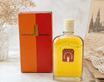 May include: A vintage perfume bottle with a yellow liquid and a cream-colored cap. The bottle is next to a red and orange box with Cyrillic text. The label on the bottle and box reads "KPACHAЯ MOCKBA".