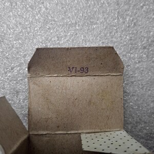 May include: Close-up of a small, open cardboard box with the text "VI-93" printed in purple. The box is a light brown color and has a textured surface. The background is a shiny, silver-gray material.