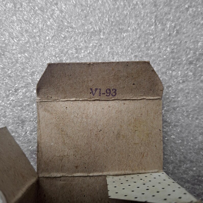 May include: Close-up of a small, open cardboard box with the text "VI-93" printed in purple. The box is a light brown color and has a textured surface. The background is a shiny, silver-gray material.