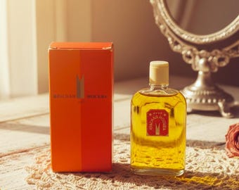May include: A vintage perfume bottle with a yellow liquid and a cream-colored cap, next to a red and orange box. The box has the words "КРАСНАЯ МОСКВА" printed on it. The perfume bottle has a red label with the same text.
