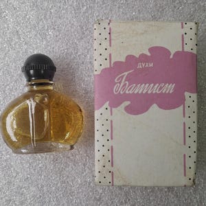 May include: A vintage perfume bottle with a black cap and a clear, rounded glass body filled with a golden liquid. The bottle sits next to a rectangular box with the word "Baptist" in cursive on a pink cloud-shaped design.