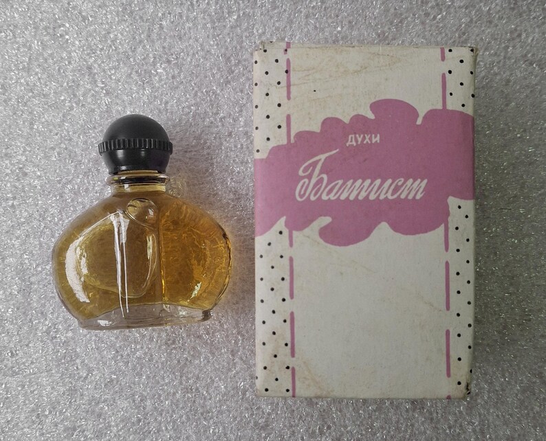 May include: A vintage perfume bottle with a black cap and a clear, rounded glass body filled with a golden liquid. The bottle sits next to a rectangular box with the word "Baptist" in cursive on a pink cloud-shaped design.