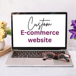 May include: A laptop computer with a white screen displaying the text "Custom E-commerce website". The laptop is on a white surface with a pair of glasses and a book.