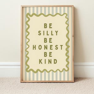 Op de afbeelding: Een ingelijste print met de tekst "BE SILLY BE HONEST BE KIND" in groene letters. De achtergrond heeft verticale strepen in lichtgroen en wit. De lijst is lichtbruin.