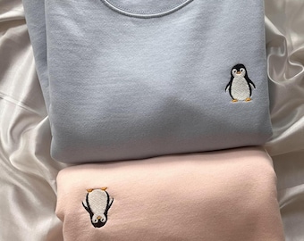 Könnte beinhalten: Zwei Sweatshirts mit Pinguin-Stickerei. Das obere Sweatshirt ist hellblau mit einem nach vorne blickenden Pinguin. Das untere Sweatshirt ist rosa mit einem nach hinten blickenden Pinguin. Beide Sweatshirts haben ein Etikett mit der Aufschrift "NEL".