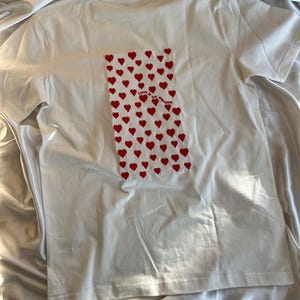May include: White t-shirt with a red heart pattern and the text "Love is for everyone" printed on the back.