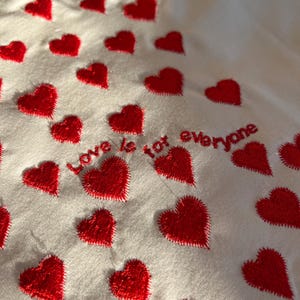 May include: White fabric with red embroidered hearts and the text "Love is not everyone".