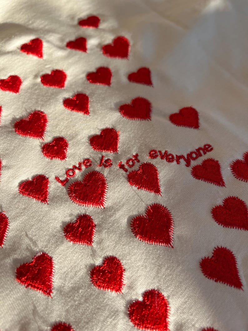 May include: White fabric with red embroidered hearts and the text "Love is not everyone".