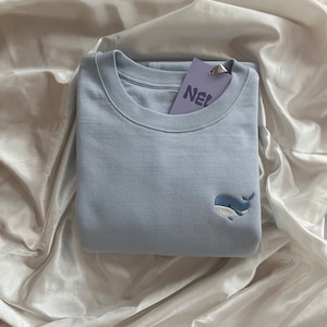Könnte beinhalten: Hellblaues Sweatshirt mit Rundhalsausschnitt und kleinem, gesticktem Wal-Design in Blau und Weiß. Ein lila Etikett mit dem Wort "NEW" ist am gefalteten Kleidungsstück befestigt.