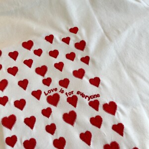 May include: White t-shirt with red embroidered hearts and the text "Love is for everyone" on the back.