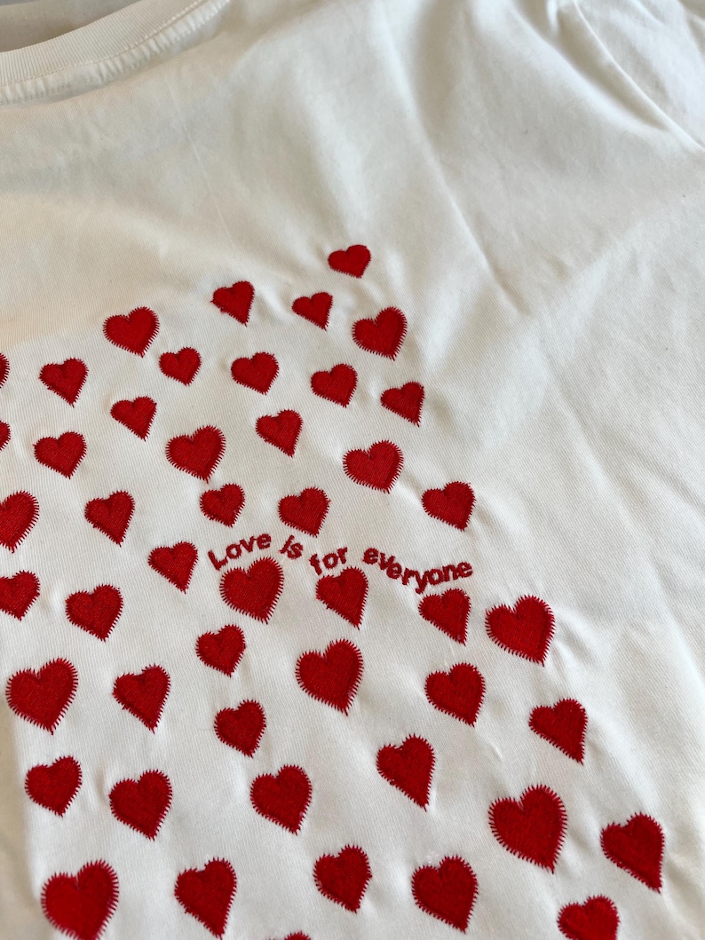 May include: White t-shirt with red embroidered hearts and the text "Love is for everyone" on the back.