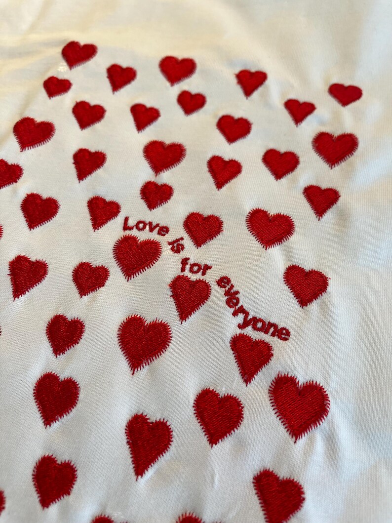 May include: White t-shirt with red embroidered hearts and the text "Love is for everyone".
