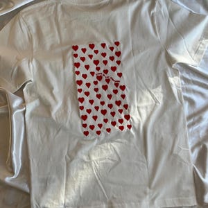 May include: White t-shirt with a red heart pattern and the text "Love for everyone" on the back.