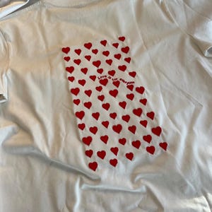 May include: White t-shirt with a red heart pattern and the text "Love is for everyone" on the back.