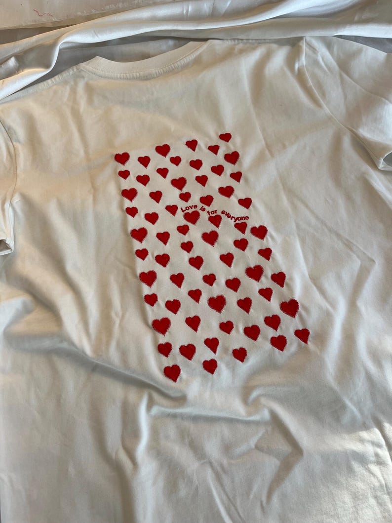 May include: White t-shirt with a red heart pattern and the text "Love is for everyone" on the back.