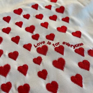 May include: White t-shirt with red embroidered hearts and the text "Love is for everyone" in a red embroidered font.