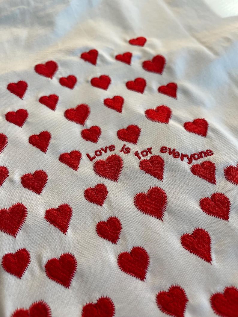 May include: White t-shirt with red embroidered hearts and the text "Love is for everyone" in a red embroidered font.
